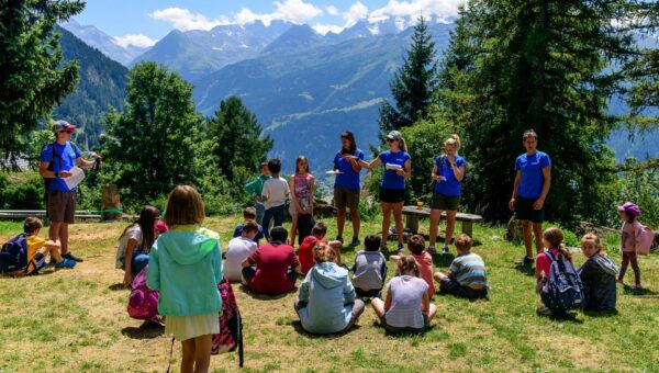 Children seated on a grassy hillside, listening to instructors in blue shirts with mountains in the background.